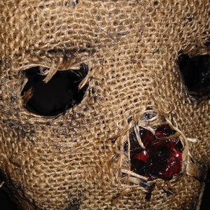 Puede incluir: Primer plano de una m&aacute;scara de arpillera con grandes agujeros para los ojos y la boca. La m&aacute;scara tiene una superficie rugosa y texturizada con manchas oscuras y una mancha roja oscura cerca de la boca. Accesorio de disfraz de Halloween.