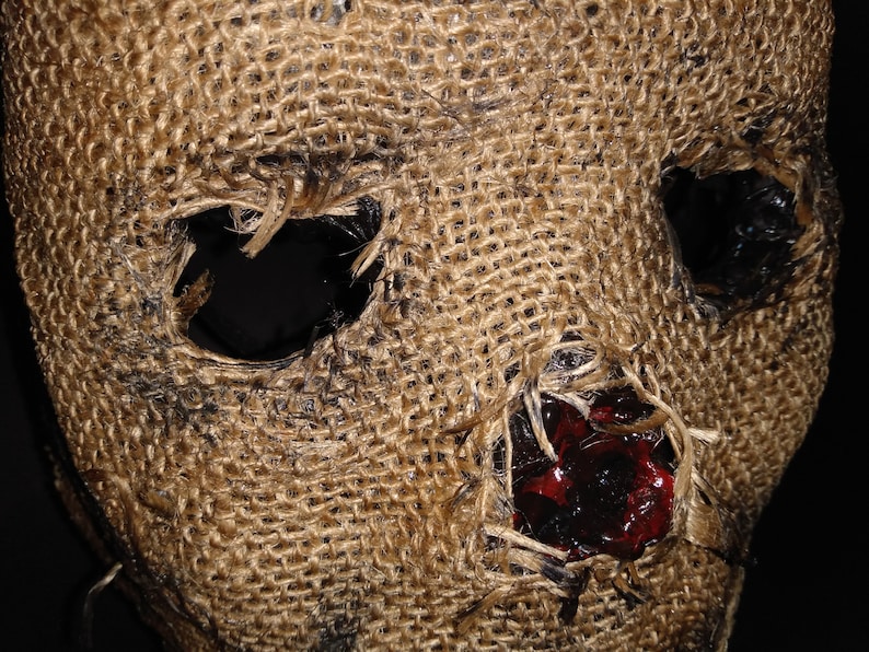 Puede incluir: Primer plano de una m&aacute;scara de arpillera con grandes agujeros para los ojos y la boca. La m&aacute;scara tiene una superficie rugosa y texturizada con manchas oscuras y una mancha roja oscura cerca de la boca. Accesorio de disfraz de Halloween.