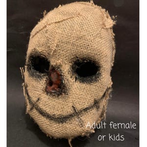 Puede incluir: Una m&aacute;scara de arpillera con cuencas de ojos negras y una sonrisa cosida. La m&aacute;scara tiene un aspecto desgastado con costuras visibles y una nariz marr&oacute;n rojiza. El texto "Adult female or kids" es visible en la parte inferior de la imagen. Esta m&aacute;scara es adecuada para Halloween o fiestas de disfraces.