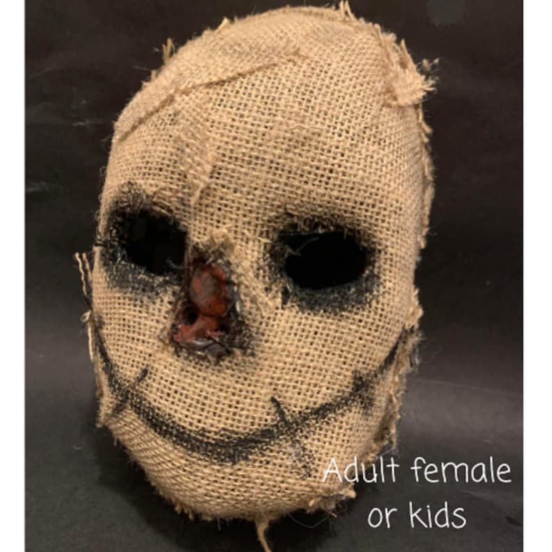Puede incluir: Una m&aacute;scara de arpillera con cuencas de ojos negras y una sonrisa cosida. La m&aacute;scara tiene un aspecto desgastado con costuras visibles y una nariz marr&oacute;n rojiza. El texto "Adult female or kids" es visible en la parte inferior de la imagen. Esta m&aacute;scara es adecuada para Halloween o fiestas de disfraces.