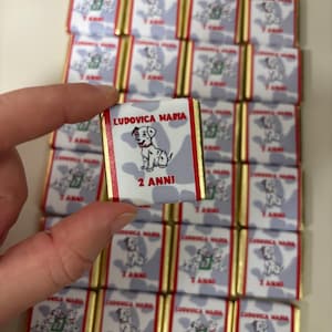 Könnte beinhalten: Kleine Schokoladenriegel mit einer Illustration eines Dalmatinerwelpen. Die Riegel haben goldene und rote Akzente und den Text "LUDOVICA MARIA 2 ANNI". Im Hintergrund sind weitere Riegel gestapelt.