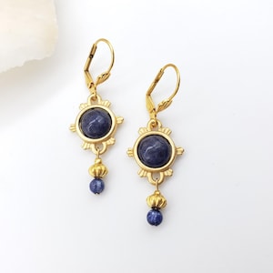 May include: A pair of gold-colored earrings with a dark blue, round stone center. The earrings have a sunburst design around the stone and a small blue bead hanging below. The earrings are on a white background.