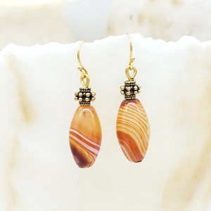 May include: A pair of gold dangle earrings with oval-shaped agate stones. The stones are a mix of brown, white, and orange with a striped pattern. The earrings have a gold bead at the top.