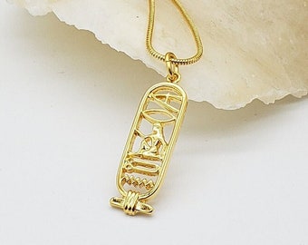 May include: A gold-colored pendant necklace featuring an Egyptian cartouche design. The pendant is suspended from a delicate gold chain. The necklace is displayed on a white surface with a light-colored stone in the background.