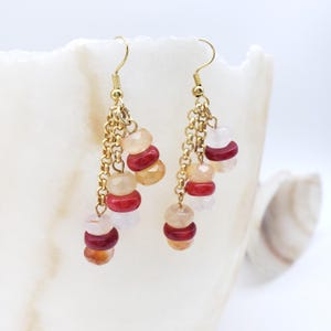 May include: Dangling earrings with gold-colored hooks and chains. Each earring features a cluster of colorful beads in shades of red, peach, and white. The earrings are set against a white background.