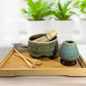 May include: A bamboo tray with a green and brown ceramic matcha bowl, a bamboo whisk, a metal strainer, a bamboo spoon, and a green and brown ceramic teacup. The tray has the text "Kit Matcha Co." printed on it.