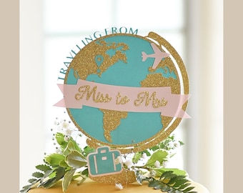 May include: A turquoise and gold glitter globe cake topper with a pink banner that says "Miss to Mrs." and a small airplane on top. The globe is surrounded by greenery.