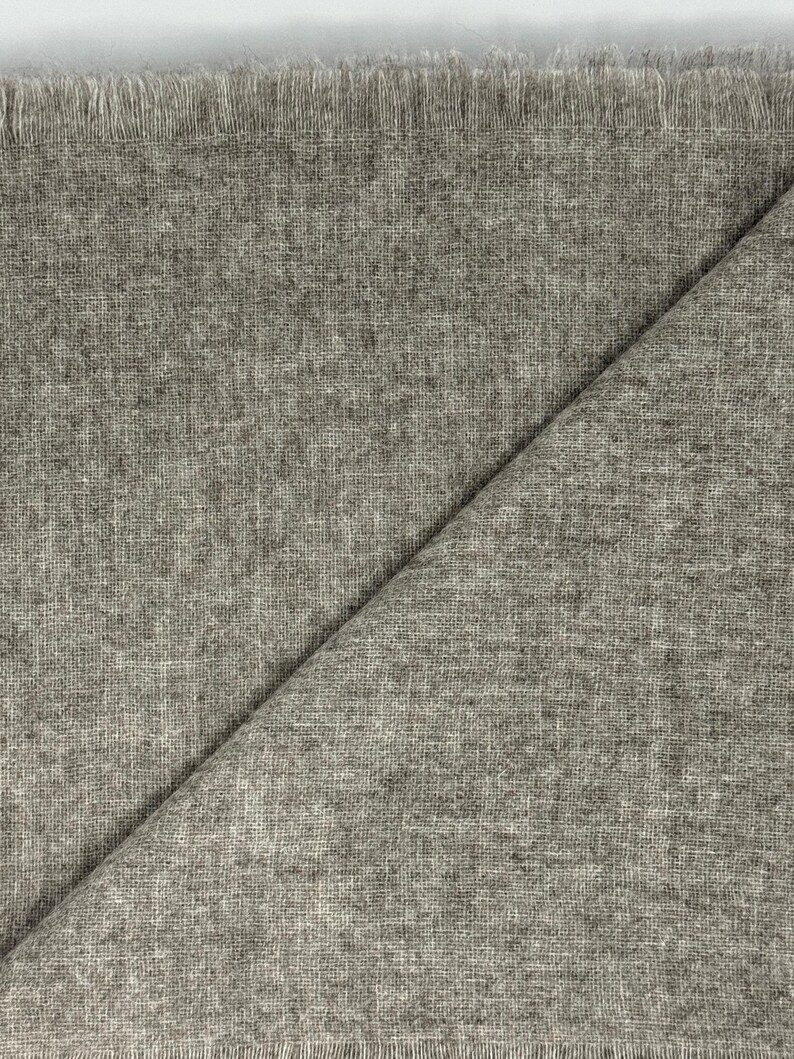 Puede incluir: Primer plano de una tela gris claro con un borde deshilachado. La tela est&aacute; doblada diagonalmente, revelando un tejido texturizado. El material parece suave y ligero, adecuado para bufandas o chales.