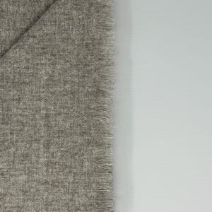 Puede incluir: Una secci&oacute;n de tela gris con un tejido texturizado y un borde con flecos. La tela est&aacute; doblada, revelando una forma triangular. El fondo es una superficie blanca lisa.