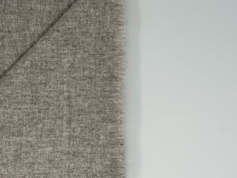 Puede incluir: Una secci&oacute;n de tela gris con un tejido texturizado y un borde con flecos. La tela est&aacute; doblada, revelando una forma triangular. El fondo es una superficie blanca lisa.