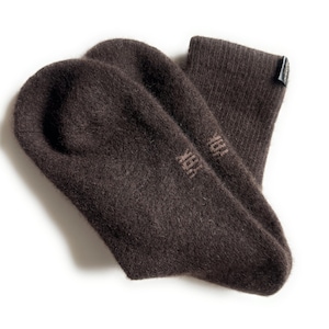 May include: A pair of dark brown socks with a soft, textured appearance. The socks are made of a thick material and have the word "YAK" printed on the foot. A small tag is attached to the top of one sock.