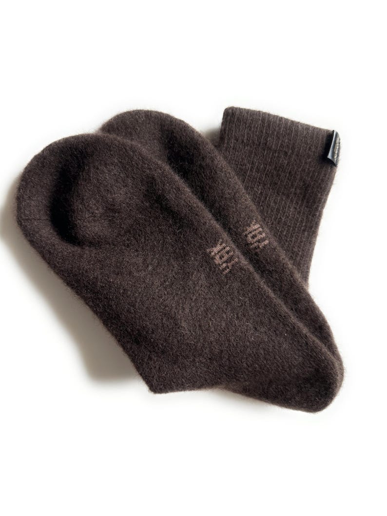 May include: A pair of dark brown socks with a soft, textured appearance. The socks are made of a thick material and have the word "YAK" printed on the foot. A small tag is attached to the top of one sock.