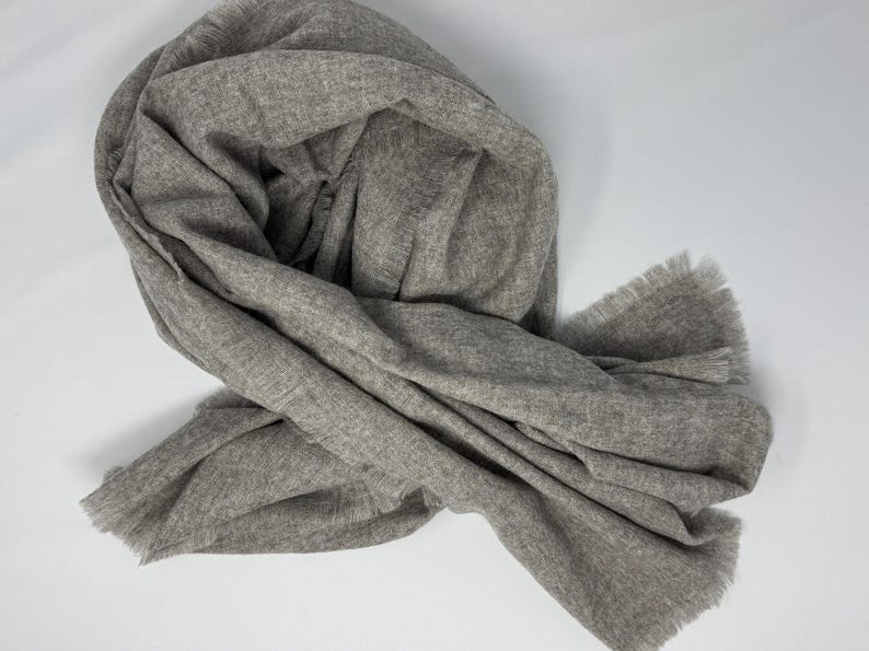 Puede incluir: Bufanda gris claro con borde deshilachado, drapeada y doblada sobre una superficie blanca. La bufanda parece estar hecha de un material suave y tejido. El color neutro y el dise&ntilde;o sencillo sugieren un accesorio vers&aacute;til.