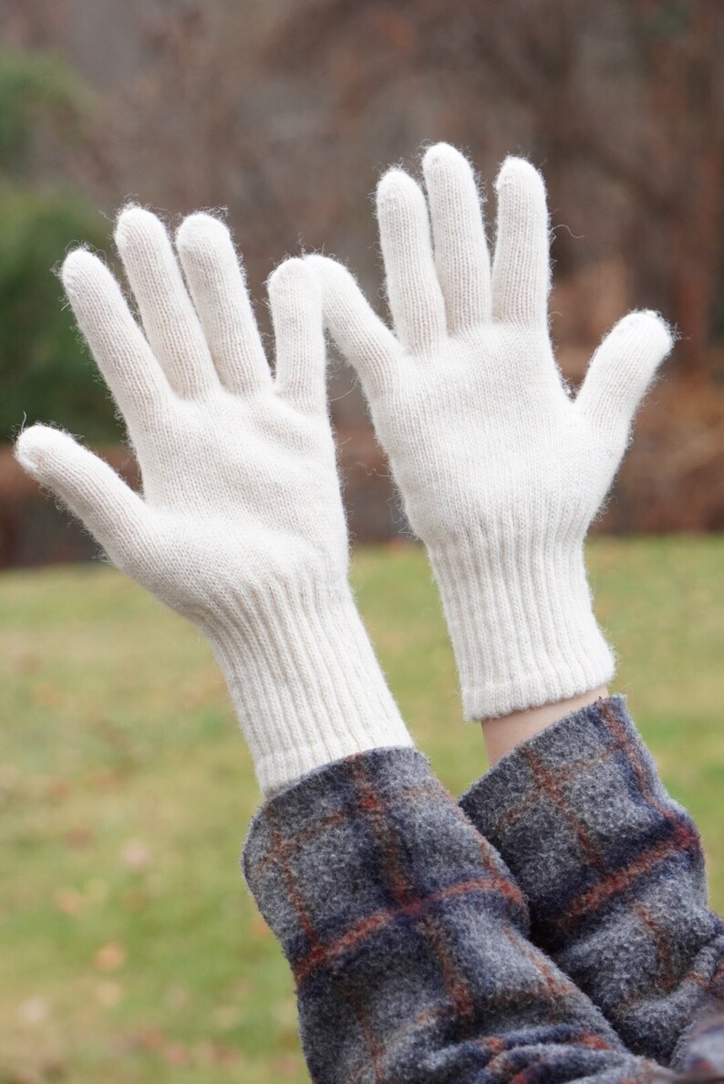 May include: A pair of white knitted gloves with a ribbed cuff. The gloves are held up in the air with the fingers spread out.