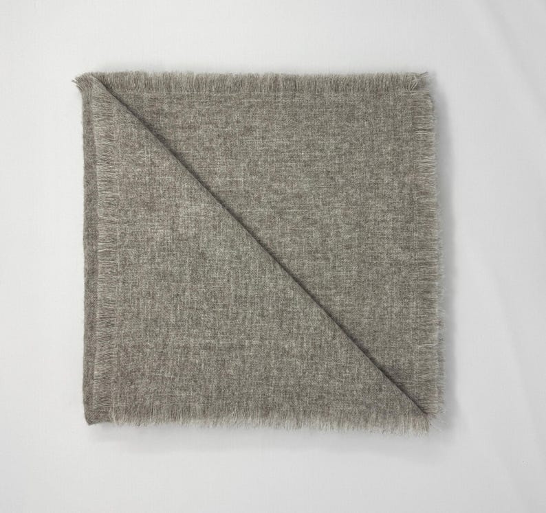 Puede incluir: Una manta gris claro doblada con un borde con flecos. La manta est&aacute; doblada diagonalmente, creando una forma triangular. La tela parece suave y texturizada, adecuada para la decoraci&oacute;n del hogar.