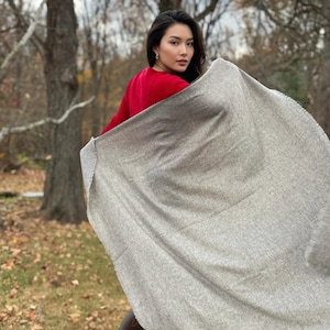 Puede incluir: Una persona con un su&eacute;ter rojo y botas marrones sostiene una bufanda grande de color gris claro al aire libre. La bufanda tiene un borde con flecos y parece estar hecha de un material suave y tejido. El fondo presenta &aacute;rboles y follaje oto&ntilde;al.