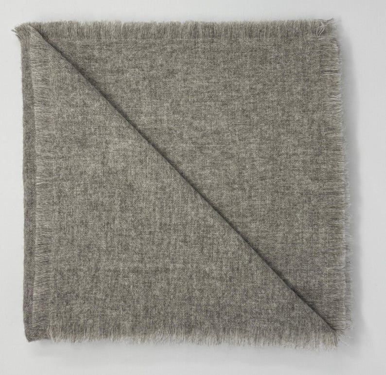 Puede incluir: Una manta cuadrada plegada, de color gris claro, con un borde con flecos. La manta est&aacute; doblada diagonalmente, creando una forma triangular. La tela parece suave y texturizada, adecuada para la decoraci&oacute;n del hogar.