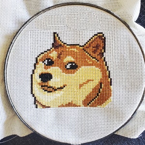 May include: Cross-stitch embroidery of a Shiba Inu dog's face with a brown and white fur pattern. The dog is looking to the right with a slight smile.