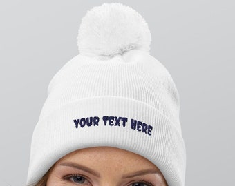 Può includere: Berretto di lana bianco con un pon pon bianco in cima. Il berretto ha il testo "YOUR TEXT HERE" ricamato in blu scuro.