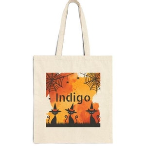 Puede incluir: Un bolso tote de color crema con un diseño de Halloween. El diseño presenta siluetas de gatos negros con sombreros de bruja, telarañas y la palabra "Indigo" en naranja. El fondo es un lavado de acuarela naranja y rojo.