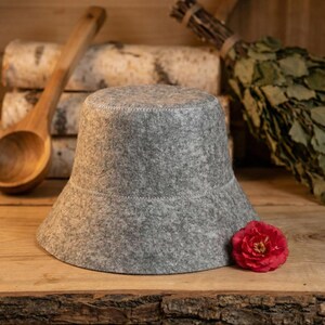 K&ouml;nnte beinhalten: Ein grauer Sauna-Filzhut mit breiter Krempe und wei&szlig;en N&auml;hten. Im Hintergrund befinden sich eine Holzkelle, Birkenst&auml;mme und eine rote Blume. Der Hut soll den Kopf vor Hitze sch&uuml;tzen.