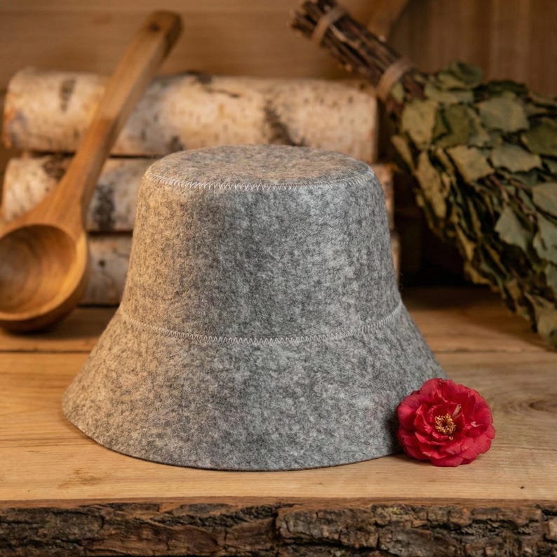 K&ouml;nnte beinhalten: Ein grauer Sauna-Filzhut mit breiter Krempe und wei&szlig;en N&auml;hten. Im Hintergrund befinden sich eine Holzkelle, Birkenst&auml;mme und eine rote Blume. Der Hut soll den Kopf vor Hitze sch&uuml;tzen.