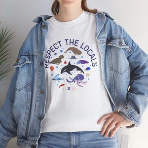 Puede incluir: Camiseta blanca con un diseño circular de animales marinos, como una orca, un pulpo y peces. El texto azul marino dice "Respect the Locals". La modelo lleva una chaqueta vaquera azul claro. El diseño es una ilustración colorida.