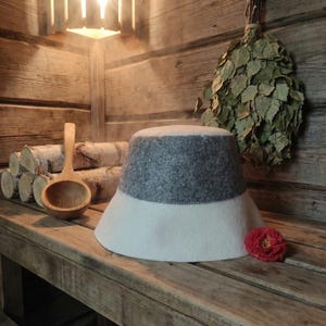 Puede incluir: Un sombrero de sauna de dos tonos en gris y blanco, con ala ancha. Una cuchara de madera y troncos de abedul son visibles sobre una superficie de madera. Una flor roja y un manojo de hojas de abedul cuelgan en el fondo. El sombrero protege la cabeza del calor.