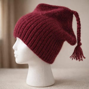 May include: A burgundy knitted hat with a distinctive pointed shape and a braided tassel. The hat is displayed on a white mannequin head, highlighting its texture and design. The hat's colour is a deep, rich red.