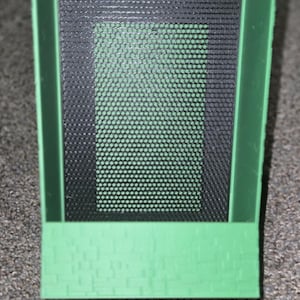 May include: A green rectangular object with a black mesh screen. The object has a brick-like pattern on the bottom. The object is sitting on a gray surface.