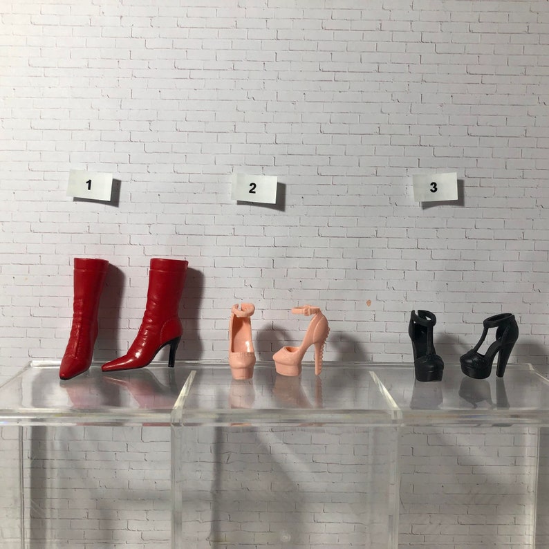 Puede incluir: Tres pares de zapatos de mu&ntilde;eca sobre un fondo de ladrillo blanco. El primer par son botas rojas, el segundo par son tacones altos rosas y el tercer par son tacones altos negros.