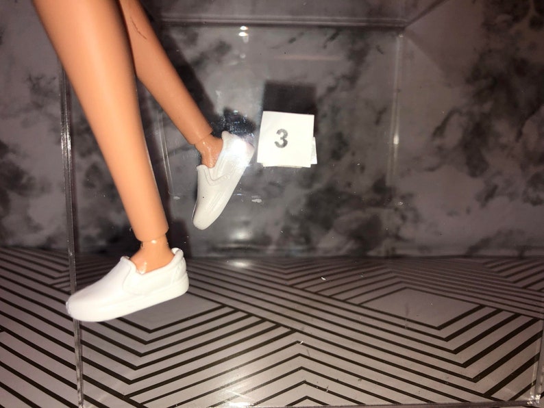 May include: A pair of white slip-on shoes with a simple design. The shoes are displayed in a clear plastic box with a number "3" written on a piece of paper.