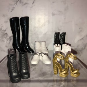 May include: A collection of eight pairs of doll shoes, including black boots, white ankle boots, white flats, and gold heeled sandals.