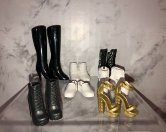May include: A collection of eight pairs of doll shoes, including black boots, white ankle boots, white flats, and gold heeled sandals.