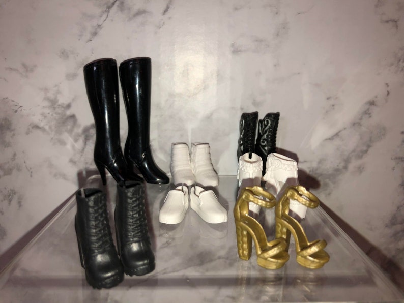 May include: A collection of eight pairs of doll shoes, including black boots, white ankle boots, white flats, and gold heeled sandals.