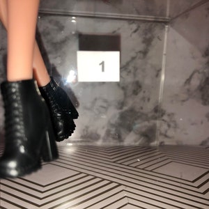 May include: A pair of black ankle boots with laces and a chunky heel. The boots are on a patterned surface with the number "1" visible in the background.