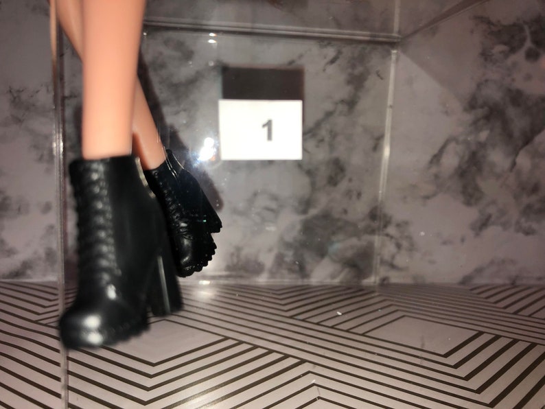 May include: A pair of black ankle boots with laces and a chunky heel. The boots are on a patterned surface with the number "1" visible in the background.