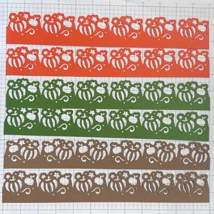 May include: Rows of die-cut paper strips in orange, green, and brown. Each strip features a repeating pumpkin design with intricate cut-out details. The strips are arranged on a grid, showcasing the decorative patterns.