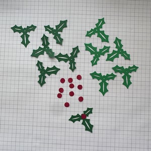 May include: A collection of green paper holly leaf cutouts and small red paper circle cutouts. There are eight green holly leaf shapes with three points each and nine small red circles. One red circle is placed in the center of one leaf.