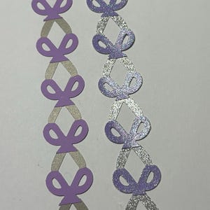 May include: Two decorative paper chains. One chain is made of lavender-colored bows connected by gray diamond shapes. The other chain has lavender bows with silver glitter diamond shapes.