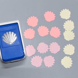 May include: A blue and white paper punch tool featuring a seashell design, accompanied by several die-cut paper seashells in shades of light pink and pale yellow, arranged in a scattered pattern on a solid gray surface.