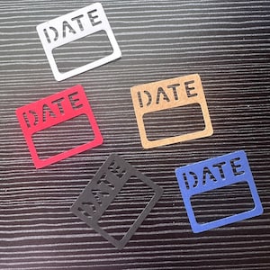 May include: Five rectangular paper labels in white, red, tan, black, and blue. Each label has the word "DATE" cut out at the top and a rectangular opening below. The labels are arranged on a dark, striped surface.