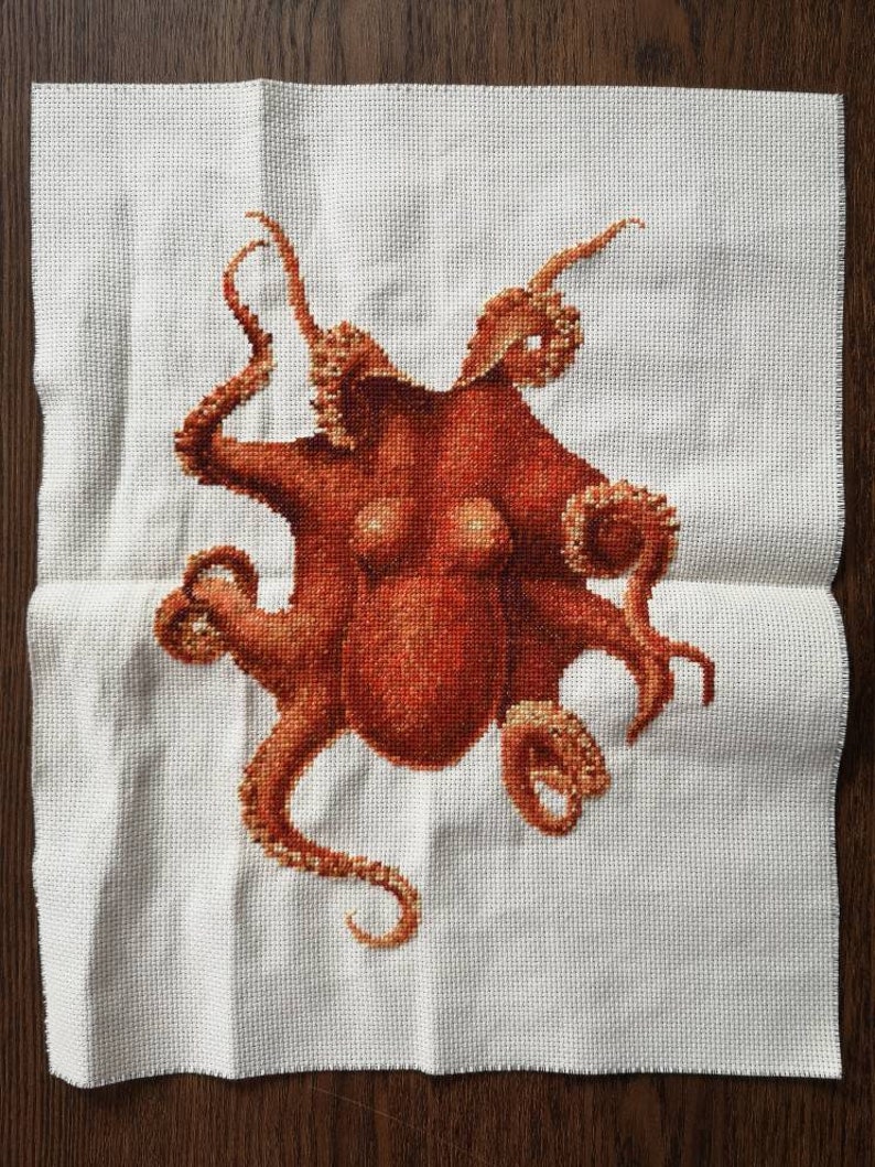 May include: A cross-stitch embroidery of an octopus with eight tentacles. The octopus is a reddish-brown color and is stitched on a white fabric.