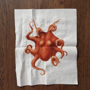 May include: A cross-stitch embroidery of an octopus with eight tentacles. The octopus is a reddish-brown color and is stitched on a white fabric.
