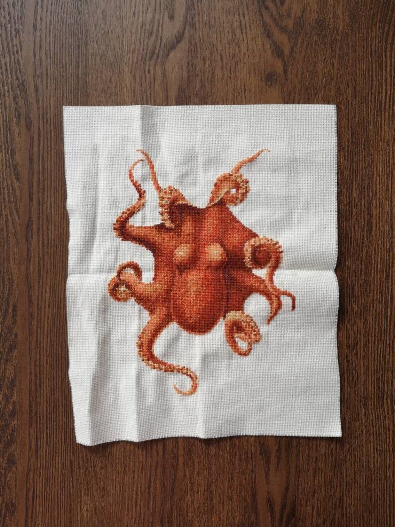 May include: A cross-stitch embroidery of an octopus with eight tentacles. The octopus is a reddish-brown color and is stitched on a white fabric.