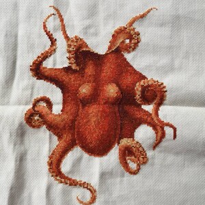 May include: A cross-stitch embroidery of an octopus with eight tentacles. The octopus is a reddish-brown color and is stitched on a white background.
