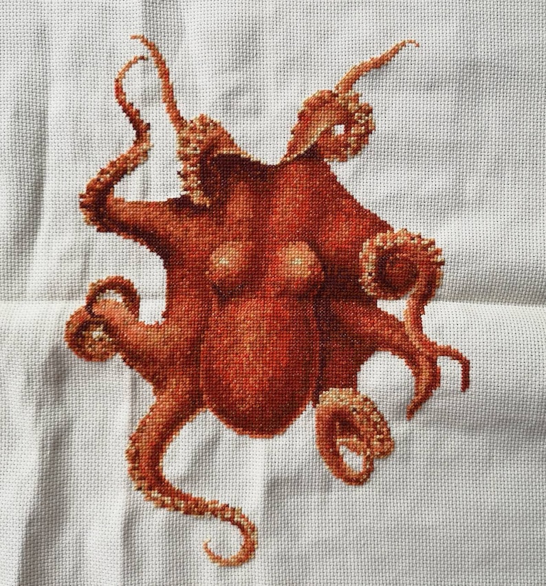 May include: A cross-stitch embroidery of an octopus with eight tentacles. The octopus is a reddish-brown color and is stitched on a white background.