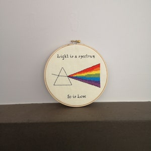 May include: A round, wooden embroidery hoop with a white fabric background. The fabric has a black and white illustration of a prism refracting light into a rainbow spectrum. The text "light is a spectrum" is above the prism and "so is love" is below the prism.