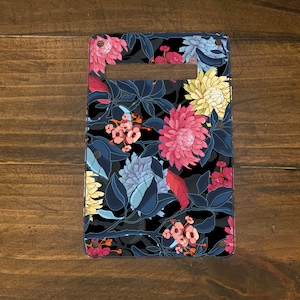 May include: Rectangular phone case with a floral pattern. The design features large pink, yellow, and blue flowers with dark blue leaves against a black background. The case has a rectangular cutout at the top.