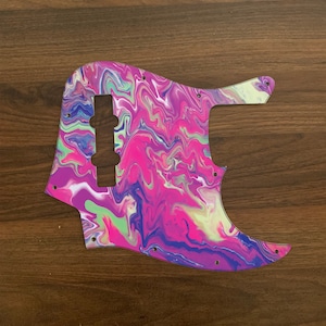 Puede incluir: Un pickguard de guitarra con un patrón abstracto de espiral en rosa, verde, azul y blanco. El pickguard está hecho de plástico y tiene un acabado liso y brillante.